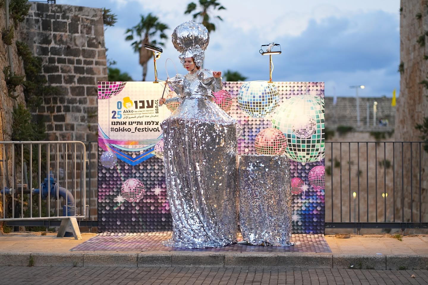 A performer in a silver costume stands by a festival poster in the street.