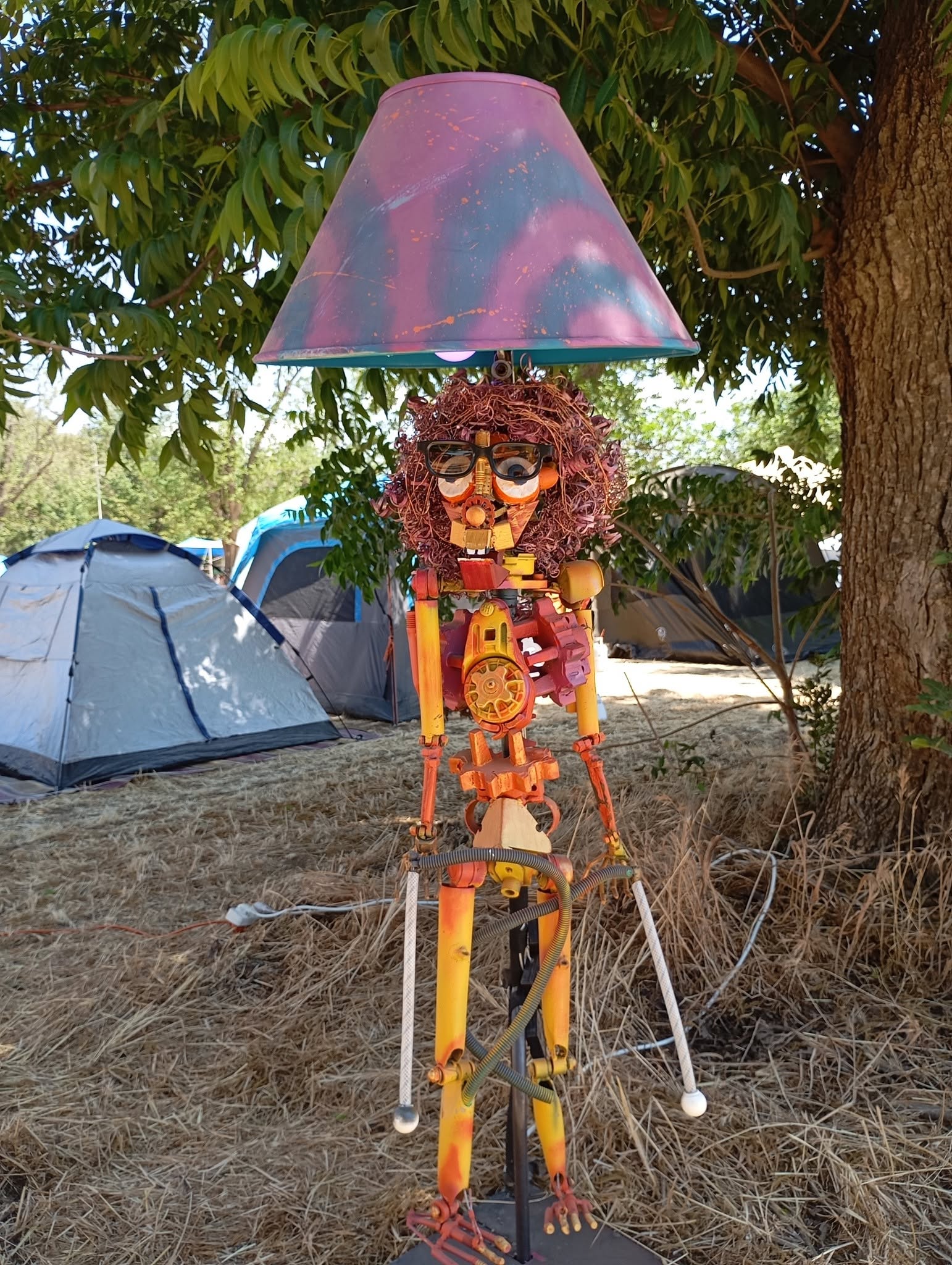 A colourful figure sculpture stands beneath a large lampshade beside tents.
