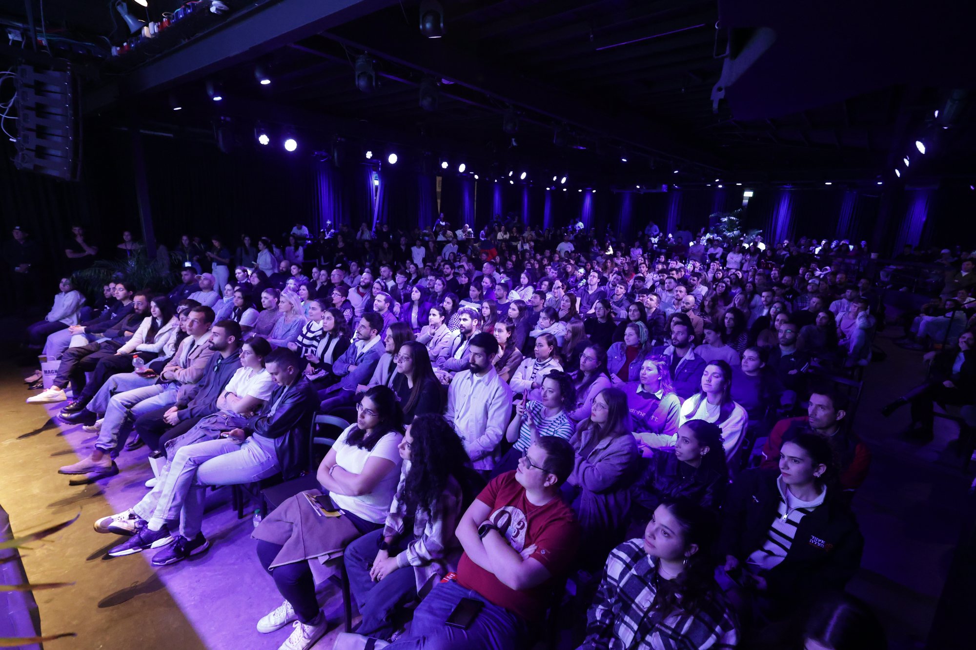 An audience sits before the conference stage under purple lighting.