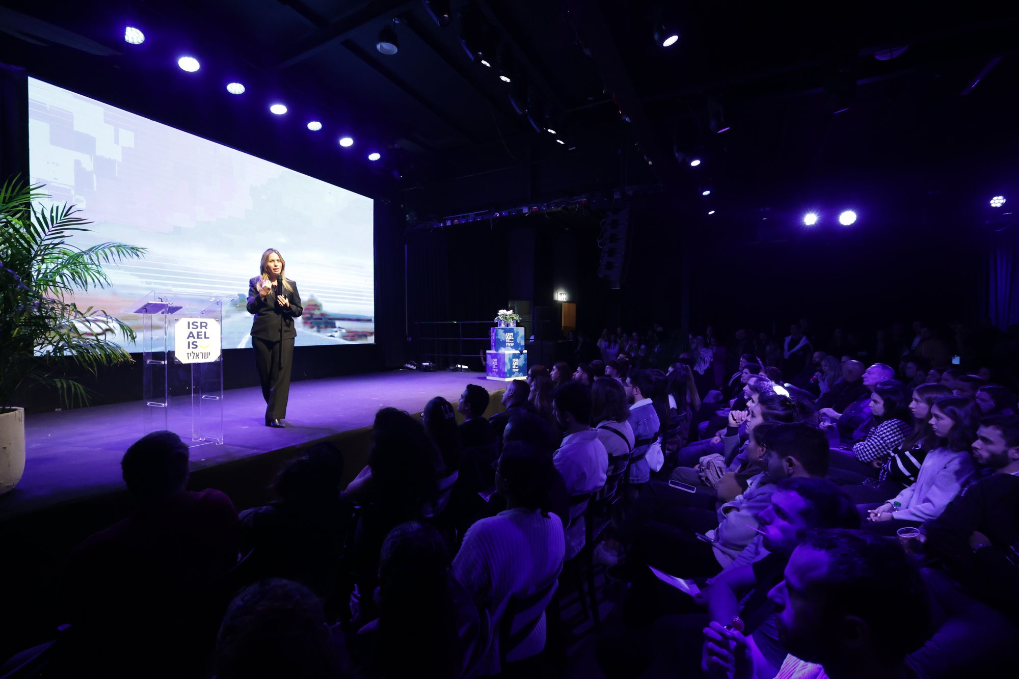 A woman speaks on stage before the audience and a presentation screen.