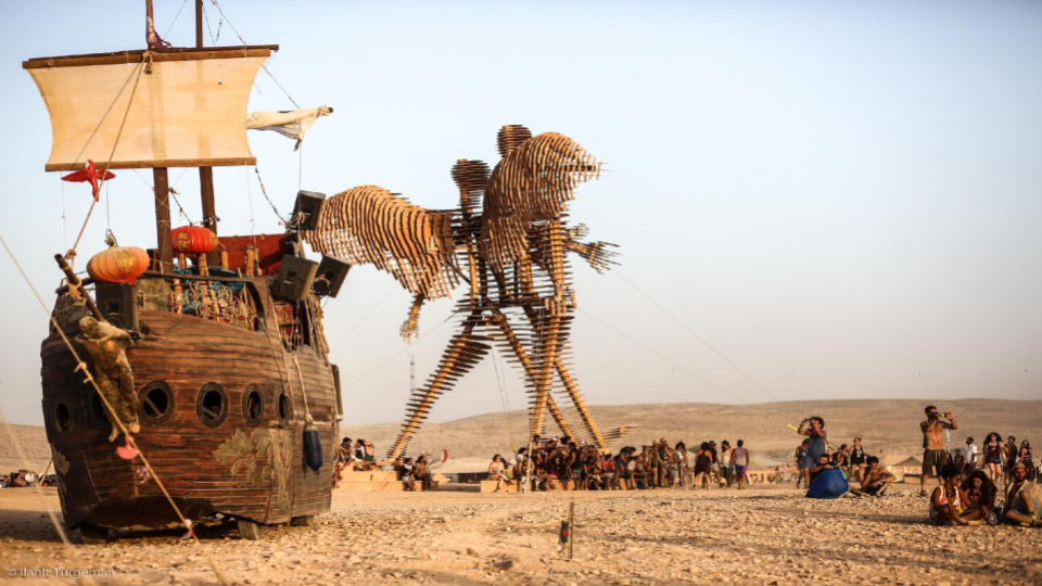 A wooden ship beside a large figure sculpture in the desert.