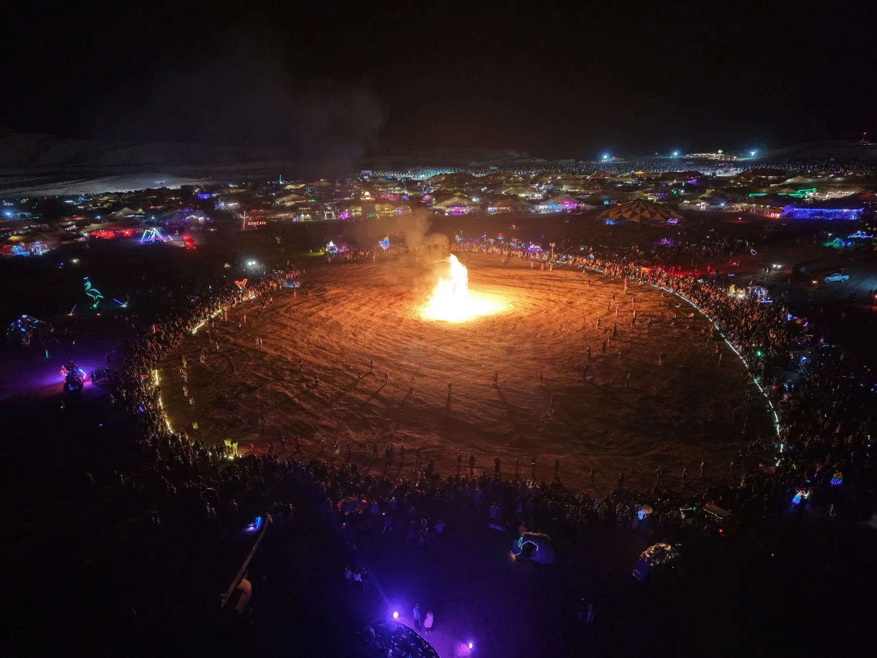 Night-time aerial view of a crowd circling a large fire.