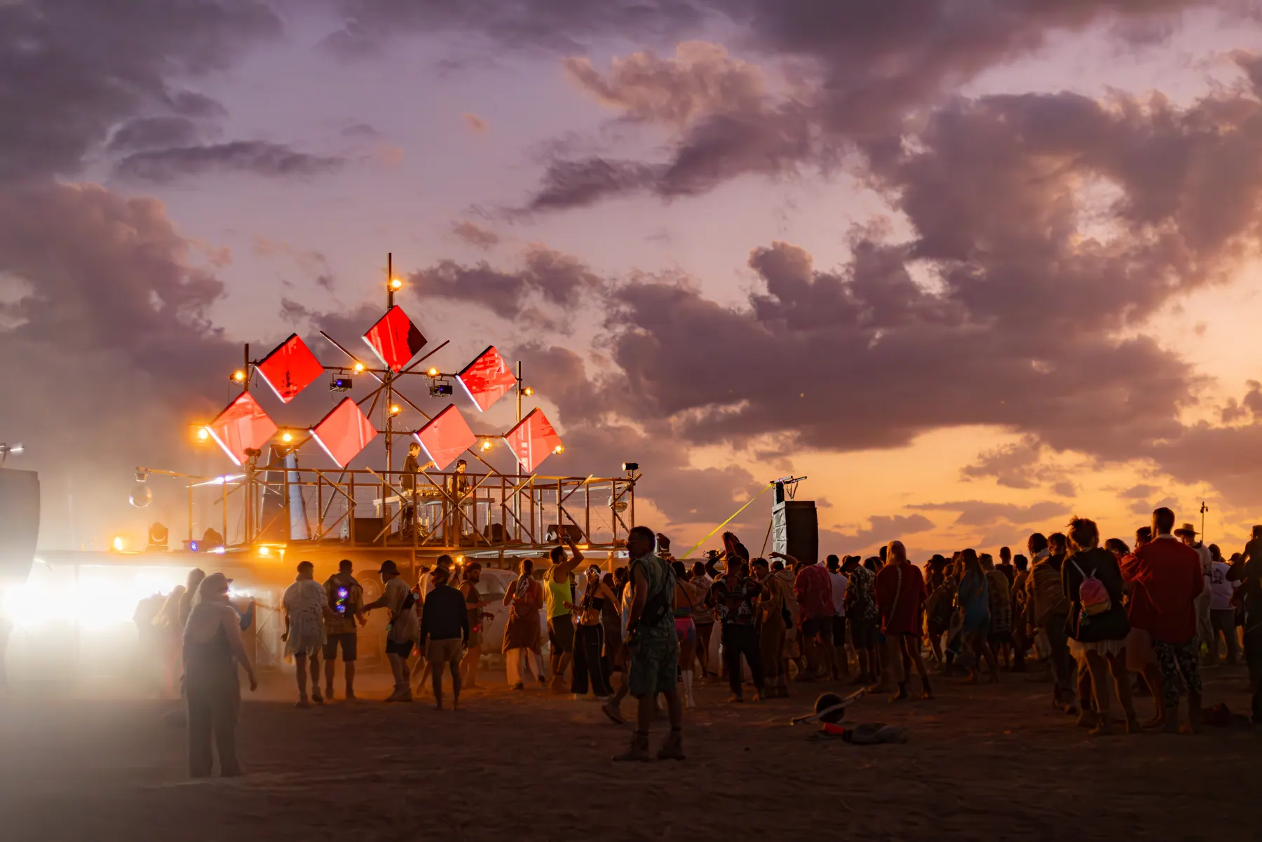 An orange-lit art structure before a crowd at dusk.