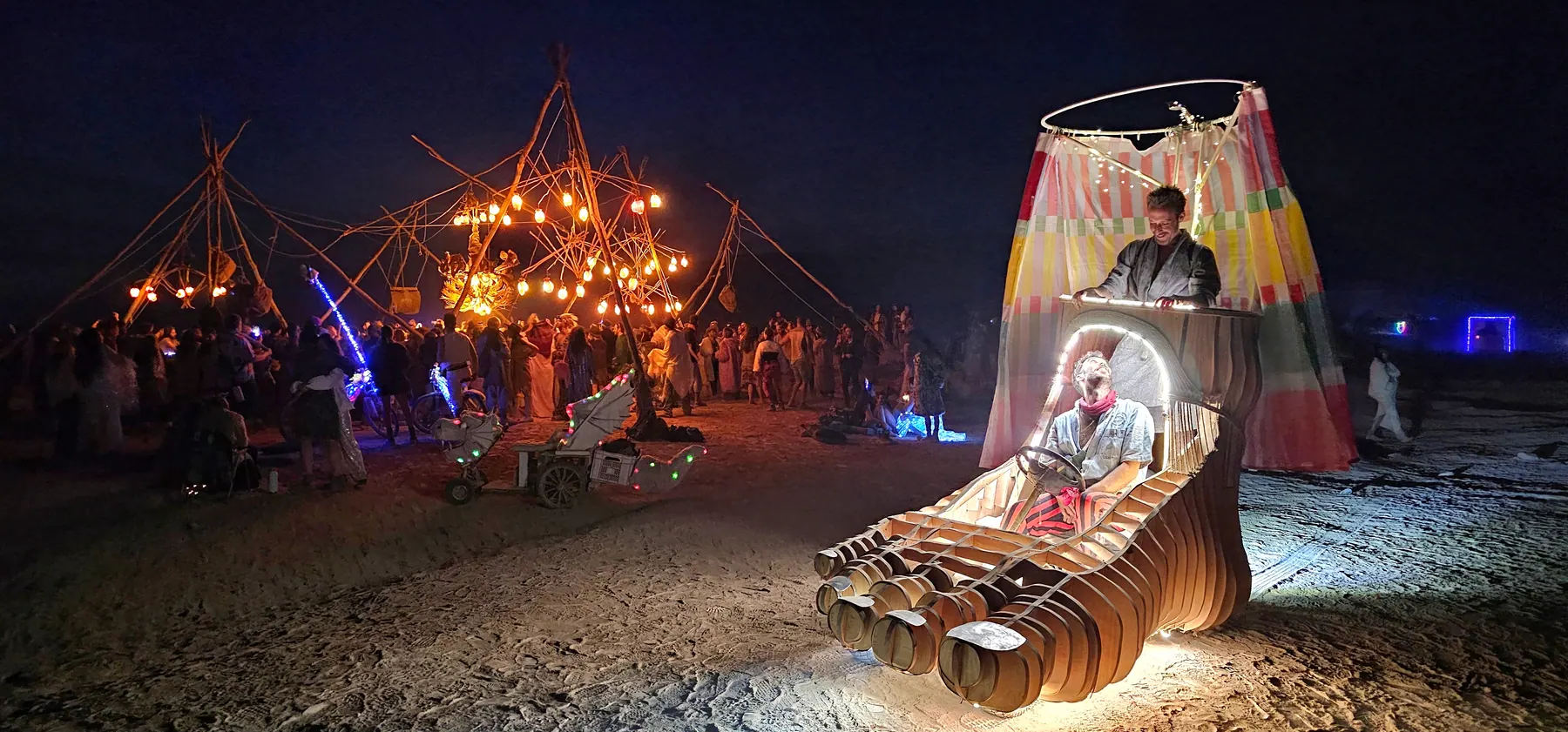 A wooden art vehicle carrying two people at a night gathering in the desert.