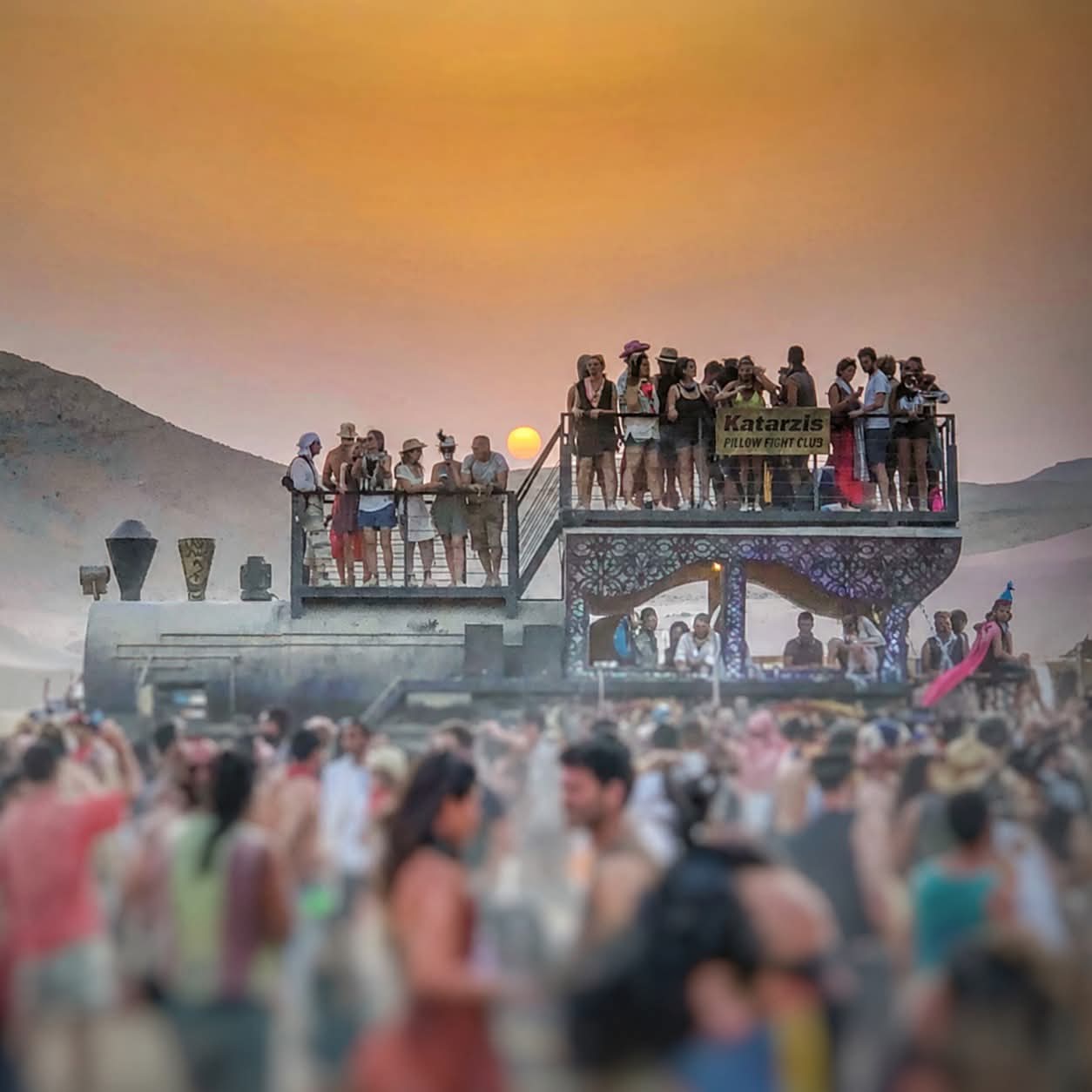 Visitors on a two-level art vehicle above a crowd at sunset.